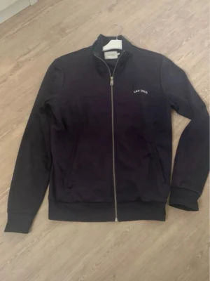 Marinblå zip hoodie från Les Deux - Snygg svart hoodie från Les Deux med dragkedja framtill och diskret logga på bröstet. Klassisk modell med ribbade muddar och ståkrage. Perfekt för en clean och stilren look. Tillverkad i mjukt material för extra komfort.