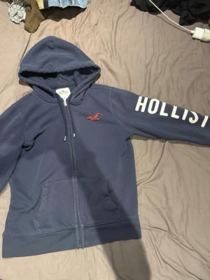 Hollister - Mörkblå zip-hoodie från Hollister med vit text på ärmen och röd broderad logga på bröstet. Hoodien har dragsko i huvan och två fickor framtill. Tillverkad i mjukt bomullsmaterial som är skönt att ha på sig.