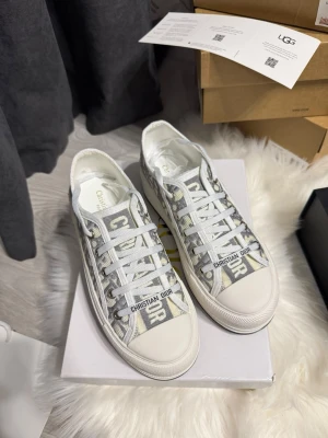 gråvita sneakers - Snygga sneakers  i grått och vitt med tydligt mönster på canvasmaterial. Skorna har vita snören och tåparti i gummi samt texten  på snörningen. Klassisk låg modell med vit sula och svart yttersula.