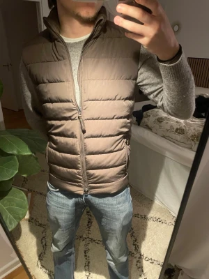 Brun dunväst med dragkedja - Snygg brun dunväst med ståkrage och horisontella sömmar. Västen har två fickor med dragkedja och stängs med en tvåvägsdragkedja framtill. Perfekt att slänga över en hoodie eller tröja för extra värme.