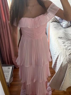 Ljusrosa offshoulder långklänning balklänning  - Romantisk ljusrosa långklänning med offshoulder och volangdetaljer. Klänningen har flera lager av plisserat tyg och en vacker axelbandslös design som ger en flowy och elegant look. Perfekt för speciella tillfällen där du vill känna dig extra fin. Helt oanvänd från lulus, lappen kvar, säljer då jag ej tyckte att jag passade i den!