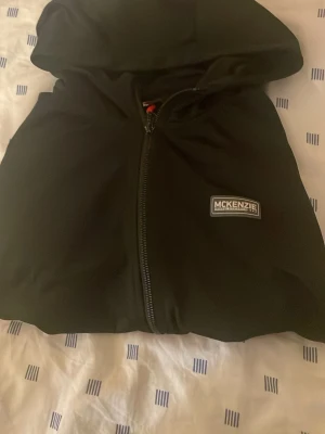 Svart hoodie med dragkedja McKenzie - Säljer en svart hoodie från McKenzie med dragkedja framtill och huva. Hoodien har en stilren look med McKenzie-logga på bröstet. Materialet känns mjukt och sportigt, perfekt för chill dagar eller när du vill ha en enkel streetwear vibe.