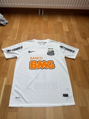 Vit Santos FC matchtröja Nike - Säljer en vit Santos FC matchtröja från Nike med Dri-FIT material. Tröjan har tryckta sponsorer som BMG och Netshoes samt Santos klubbmärke på bröstet. Perfekt för fotbollsträning eller att bära som supporter. Klassisk rund hals och korta ärmar.