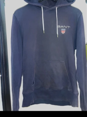 Mörkblå hoodie från GANT NY - Snygg mörkblå hoodie från GANT med vit dragsko i huvan och klassisk logga med sköld på bröstet. Hoodien har en stor magficka och ribbade muddar vid ärmslut och nederkant. Perfekt för en avslappnad och stilren look.