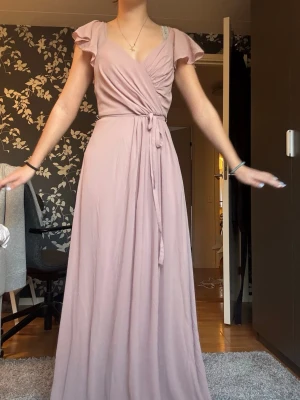 Ljusrosa omlottklänning med volangärm - Säljer en lång, ljusrosa omlottklänning med volangärmar och knytband i midjan. Klänningen har v-ringad halsringning och ett mjukt, flytande fall som ger en romantisk känsla. Perfekt för dig som vill ha en feminin och elegant look. Delvis öppen rygg