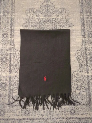 Polo Ralph Lauren halsduk - Stilren svart halsduk från Polo Ralph Lauren med klassisk röd logga broderad framtill. Halsduken har fransar i båda ändar och är tillverkad i ett mjukt material som känns lyxigt och bekvämt runt halsen.