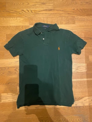Mörkgrön pikétröja från Polo Ralph Lauren - Klassisk mörkgrön pikétröja från Polo Ralph Lauren med orange broderad logga på bröstet. Tröjan har korta ärmar, krage och knappar framtill. Tillverkad i bomull för en bekväm och stilren look.