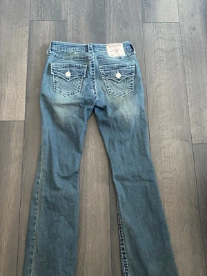True Religion jeans  - Säljer ett par blå bootcut jeans från True Religion med snygga slitningar och kontrastsömmar. Jeansen har två bakfickor med lock och dekorativa sömmar samt stora silverfärgade knappar. Klassisk femficksmodell i mjuk denim.
