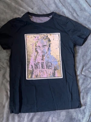 Svart t-shirt från limitato - Cool svart t-shirt från Limitato med ett stort grafiskt tryck på bröstet i lila, beige och svart. Klassisk passform och rund halsringning. Perfekt för dig som vill sticka ut med din stil.