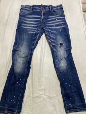 Blå distressed jeans från Dsquared2 - Säljer ett par blå jeans från Dsquared2 med slitningar, hål och färgstänk för en riktigt edgy look. Jeansen har fem fickor, raka ben och klassisk knappgylf. Materialet är denim i bomull med snygg tvätt och detaljer som sticker ut.