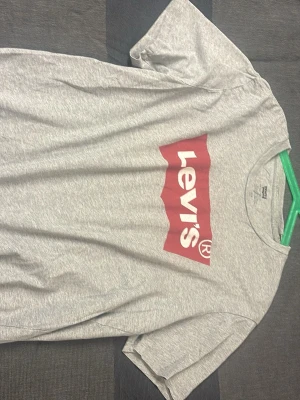 Grå Levi's t-shirt med logga - Snygg grå t-shirt från Levi's med den klassiska röda loggan tryckt framtill. T-shirten har rund halsringning och korta ärmar. Tillverkad i mjuk bomull som känns skön mot huden. Perfekt för en avslappnad och trendig stil.