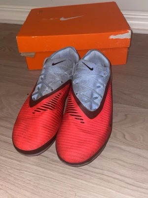 Nike phantom 6 PRO ag - Säljer ett par snygga PRO fotbollsskor från Nike i rött med blå snörning och svarta detaljer. Skorna har en modern, strömlinjeformad design och är tillverkade i syntetmaterial för bra grepp och känsla på planen. Perfekta för dig som vill sticka ut på fotbollsplanen. Pris kan diskuteras! 