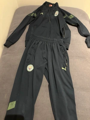 Manchester City träningsset från Puma - Snyggt svart träningsset från Puma med Manchester Citys klubbmärke på både jacka och byxor. Setet har dragkedja på jackan, elastisk midja på byxorna och detaljer i limegrönt. Perfekt för fotbollsträning eller chill. Det är storlek 176 på båda plaggen