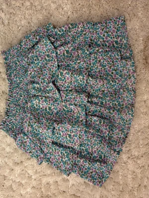 Blommig volangkjol i pastellfärger - Supersöt kort kjol med blommigt mönster i lila, blått och grönt. Kjolen har bred resår i midjan och flera volanglager som ger en luftig och lekfull känsla. Perfekt för varma dagar och enkel att styla med en crop top.