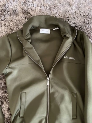 Olivgrön ziptröja från Les Deux - Snygg olivgrön ziptröja från Les Deux med hög krage och dragkedja framtill. Tröjan har två fickor och diskret Les Deux-logga på bröstet. Tillverkad i ett mjukt och stretchigt material som ger en clean och modern look.
