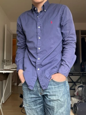 Blå skjorta från Ralph Lauren - Klassisk blå skjorta från Ralph Lauren med button-down krage och röd broderad logga på bröstet. Skjortan har långa ärmar, vita knappar och är tillverkad i mjuk bomull. Perfekt till jeans eller chinos för en clean och stilren look.