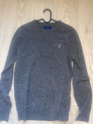Grå stickad tröja från Gant - Mysig grå stickad tröja från Gant med rund halsringning och diskret logga broderad på bröstet. Tröjan har långa ärmar och ribbade muddar vid ärmslut och nederkant. Perfekt för lager på lager och enkel att matcha till olika outfits.