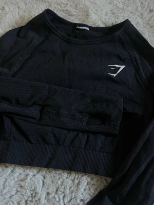 Crop top från Gymshark - Snygg svart långärmad crop top från Gymshark med tumhål i ärmarna och ventilerande meshdetaljer. Tight passform och ribbad nederkant, perfekt för träning eller gymmet. Tillverkad i stretchigt funktionsmaterial med Gymshark-logga på bröstet.