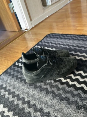 Svarta Adidas Gazelle sneakers - Säljer ett par svarta Adidas Gazelle sneakers med klassiska tre ränder på sidan. Skorna har ovandel i mocka och detaljer i läder, samt svart sula. Perfekta för dig som gillar en clean och tidlös stil.