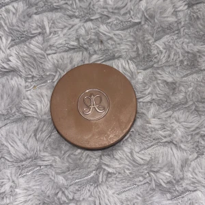 Anastasia Cream Bronzer Caramel - Säljer en rund cream bronzer från Anastasia Beverly Hills i färgen Caramel. Förpackningen är brun med märkets logga på locket och innehållet är en krämig bronzer som ger en naturlig solkysst look. Perfekt för att skulptera och värma upp ansiktet.