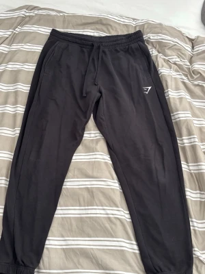 Svarta mjukisbyxor från Gymshark - Säljer ett par svarta mjukisbyxor från Gymshark med dragsko i midjan och elastiska muddar vid bensluten. Byxorna har fickor på sidorna och en diskret Gymshark-logga på vänster lår. Perfekta för chill eller träning. 