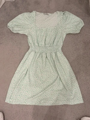 Ljusgrön blommig klänning med puffärm - Söt ljusgrön klänning med vita blommor, puffärmar och smock i midjan. Klänningen har en fyrkantig ringning och kort kjol. Materialet känns lätt, mjukt och luftigt. Klänningen är ifrån XSIDE i storlek S. Använt 1 gång :) 