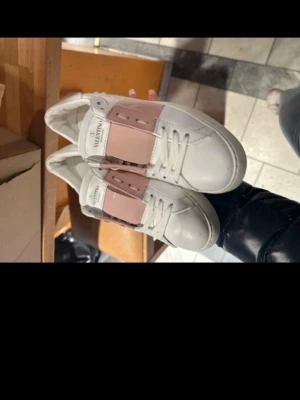 Valentino vita sneakers med rosa detalj - Snygga vita sneakers från Valentino med bred rosa rem över snörningen och logga på plösen. Skorna har klassisk låg profil och är tillverkade i slätt läder med vita skosnören. Perfekta för dig som vill ha en stilren men ändå unik look.