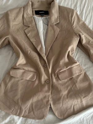 Beige kavaj från Vero Moda - Stilren beige kavaj från Vero Moda med två knappar framtill och klassisk krage. Kavajen har två fickor med lock och är helfodrad med randigt innerfoder. Perfekt för en snygg och uppklädd look.