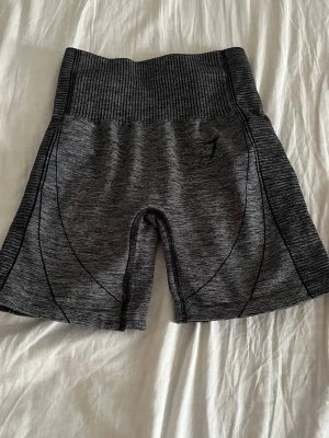 Mörkgrå träningstights shorts - Säljer ett par mörkgrå träningstights shorts med hög midja och ribbad linning. De är tillverkade i ett stretchigt syntetmaterial och har en tight passform som sitter skönt vid träning. Snygga sömdetaljer längs benen för extra stil.