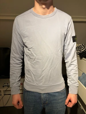 Ljusgrå sweatshirt från Stone Island - Säljer en stilren ljusgrå sweatshirt från Stone Island med klassisk rund halsringning och ribbade muddar. Tröjan har den ikoniska Stone Island-loggan på vänster ärm och är tillverkad i mjukt bomullsmaterial.