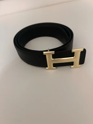 Svart bälte med gulddetalj - Snyggt svart bälte med H-spänne i guld. Bältet är gjort i skinn och har en stilren, klassisk design som passar till många olika outfits. Perfekt accessoar för att lyfta din look.