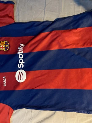 Barcelona matchtröja Lewandowski #9 - Säljer en officiell Barcelona matchtröja i rödblått med breda ränder, Spotify-logga på framsidan och Lewandowski #9 på ryggen. Tröjan har UNHCR-tryck och klubbmärke på bröstet. Perfekt för dig som älskar fotboll och vill stötta Barça.