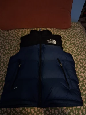 Blå och svart dunväst från The North Face - Säljer en snygg blå och svart dunväst från The North Face med dragkedja framtill och två fickor med dragkedja. Västen har hög krage och klassisk logga på bröstet och ryggen. Perfekt för lager-på-lager och riktigt varm tack vare dunfyllningen.