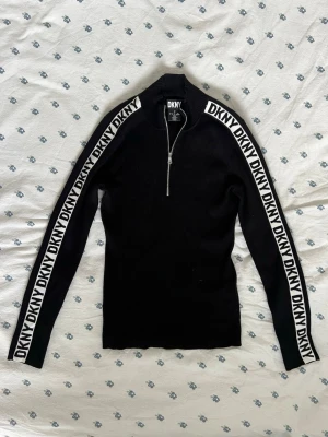 Svart vit stickad tröja från DKNY - Svart vit monogram stickad tröja från DKNY med vit logga längs båda ärmarna. Tröjan har halv dragkedja framtill, hög krage och är ribbstickad för en tight passform. Perfekt för dig som gillar sportig och trendig stil. Fitted stil men stretchiga hade passat xs-m. Måtten finns. 