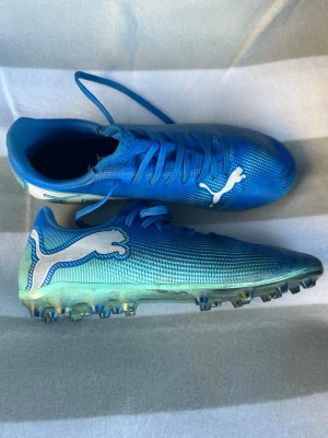 Blåa Puma fotbollsskor med dobbar - Säljer ett par blåa Puma fotbollsskor med vita detaljer och klassisk Puma-logga på sidan. Skorna har snörning och en ovandel i syntetmaterial för bra bollkontroll. Sulan har fasta dobbar som passar perfekt för spel på gräsplan. Snygg och sportig design för dig som vill sticka ut på planen.