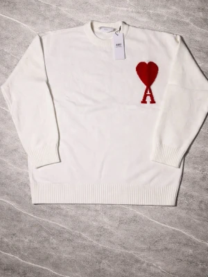 Vit stickad tröja från Ami Paris - Vit stickad tröja från Ami Paris med röd hjärtlogga och bokstaven A på bröstet. Tröjan har rund halsringning, ribbade muddar och nederkant. Perfekt för dig som gillar stilrena plagg med en twist. vet inte om den är äkta