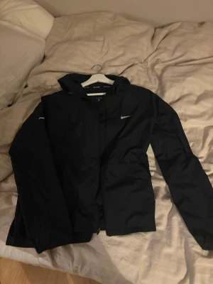 Nike windbreaker  - Svart regnjacka från Nike med minimalistisk design och diskret logga på bröstet. Jackan har huva, dragkedja framtill och är tillverkad i vattenavvisande material. Perfekt för regniga dagar och enkel att matcha med olika outfits.