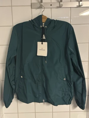 Mörkgrön vindjacka från Moncler - Snygg mörkgrön vindjacka från Moncler med huva och dragsko. Jackan har hel dragkedja framtill, två fickor med tryckknappar och diskret Moncler-logga vid halsen. Tillverkad i lätt, vattenavvisande material som passar perfekt för blåsiga dagar.
