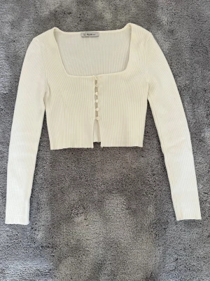Vit ribbad cropped cardigan Dilvin - Trendig vit ribbstickad cropped cardigan från Dilvin med lång ärm och fyrkantig urringning. Knäppning framtill med små knappar och mjukt stretchigt material. Perfekt att styla över ett linne eller till höga jeans.