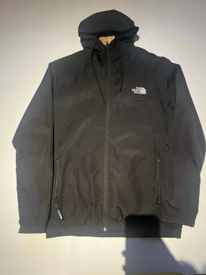  Vindjacka från The North Face - Snygg svart vindjacka från The North Face med huva och dragkedja framtill. Jackan har två sidofickor med dragkedja och justerbar dragsko i huvan. Tillverkad i ett lätt och tåligt material, perfekt för blåsiga dagar. OBS! vänster ficka är trasig!