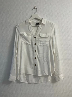 Vit skjorta med knappar från Vero Moda  - Klassisk vit skjorta från vero Moda med bruna knappar framtill och två bröstfickor. Skjortan har lång ärm och en avslappnad passform. Perfekt till jeans eller kjol för en clean look.