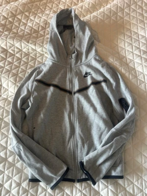 Grå zip hoodie från Nike - Säljer denna supersnygga gråa Nike techen som både passar tjejer och killar till bra pris. Passar de som är mellan 158-170cm långa. Bra till okej skick då färgen runt de svarta ränderna ändrats lite under tvättning. Pris går att diskutera 💞