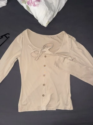 Beige topp med knyt och knappar - Snygg beige topp med långa ärmar, knytning vid halsen och knappar framtill. Perfekt för dig som gillar en enkel men ändå detaljrik stil. Passar till det mesta och är lätt att matcha med andra plagg.