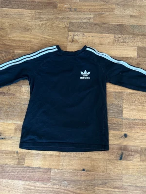 Svart Adidas sweatshirt med ränder - Svart långärmad sweatshirt från Adidas med klassiska vita ränder längs ärmarna och Adidas-logga på bröstet. Tröjan har rund hals och är tillverkad i mjukt material, perfekt för en sportig och avslappnad stil.