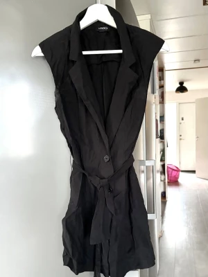 Ärmlös svart kavaj från Lindex - Stilren svart ärmlös kavaj från Lindex med klassisk krage, knäppning framtill och matchande midjebälte. Perfekt för att lyfta din outfit med en modern touch. Materialet ser ut att vara lätt och mjukt, vilket ger en snygg passform.