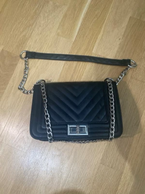 Svart quiltad axelväska med kedja - Snygg svart axelväska med quiltat mönster och silverfärgad kedjerem. Väskan har en rektangulär form, lock med metallspänne och detaljer i metall.