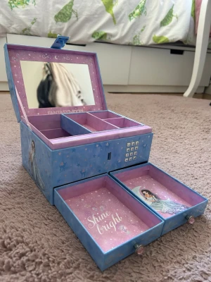 Blå smyckeskrin Fantasy Model - Snyggt smyckeskrin i blått med lila insida och spegel i locket. Utsidan har motiv på en av tjejerna ur TOPModel franchaisen. Två utdragbara lådor med texten 'Shine bright' och illustrationer. Skrinet har kodlås och flera fack för smycken. Tillverkat i hård papp. Smyckeskrinet är en utgående modell.