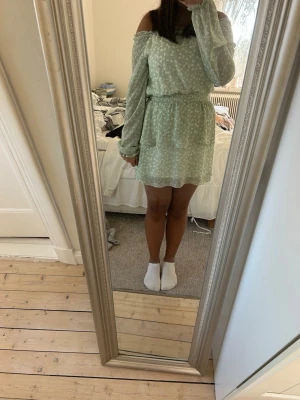 Ljusgrön offshoulder blommig klänning - Söt ljusgrön kortklänning med små vita blommor, offshoulder-modell eller inte (frivilligt) och långa ärmar. Klänningen har resår i midjan för en bekväm passform och är tillverkad i ett lätt, luftigt material. Perfekt för varma dagar och somriga tillfällen.