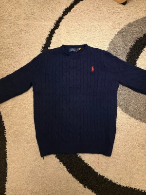 Mörkblå kabelstickad tröja Polo Ralph Lauren - Snygg mörkblå kabelstickad tröja från Polo Ralph Lauren med klassisk röd logga broderad på bröstet. Tröjan har rund halsringning och ribbade muddar vid ärmslut och nederkant. Perfekt för dig som gillar stilrena och tidlösa plagg.