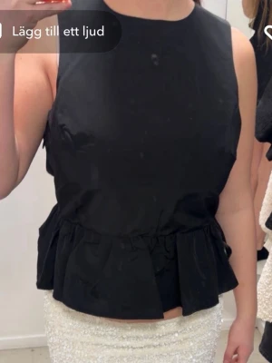 Svart ärmlös topp med rosett - Snygg svart ärmlös topp med volang nedtill och stor rosett i ryggen. Toppen har öppen rygg och är croppad, vilket ger en trendig och ungdomlig look. Perfekt att styla med kjol eller jeans för en cool outfit.